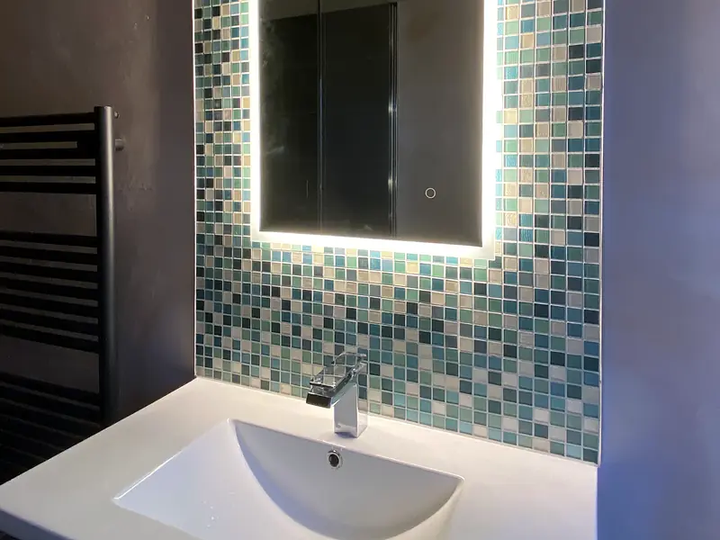 Bathroom basin and mixer tap beneath an illuminated mirror with a mosaic tile splashback